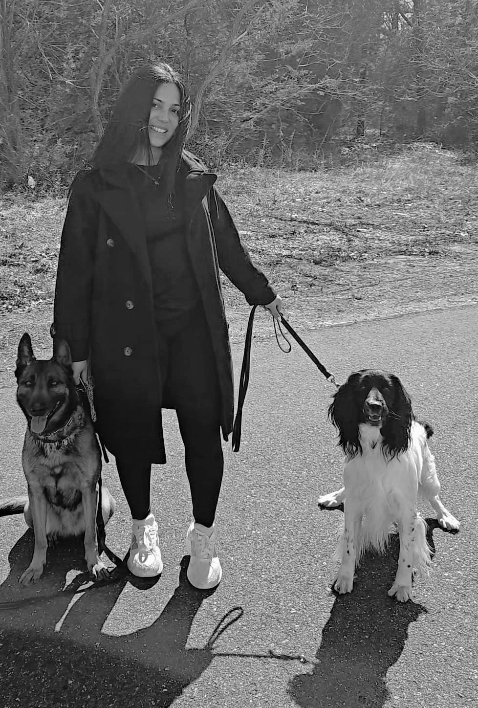 Woman in black coat walking two dogs on leashes on a tree-lined path in black and white